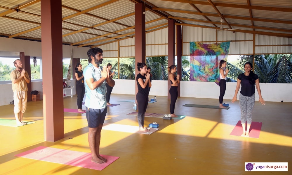 Yoga students in class with hands in anjali mudra (prayer) as the yoga teacher stands at the front of the class in samastitih (mountain pose) during yoga teacher training in Goa, India