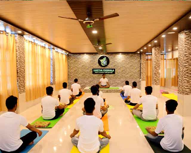 A yoga shala full of students in a seated, cross-legged posture and listening to the teacher during their yoga teacher training in Rishikesh, India.